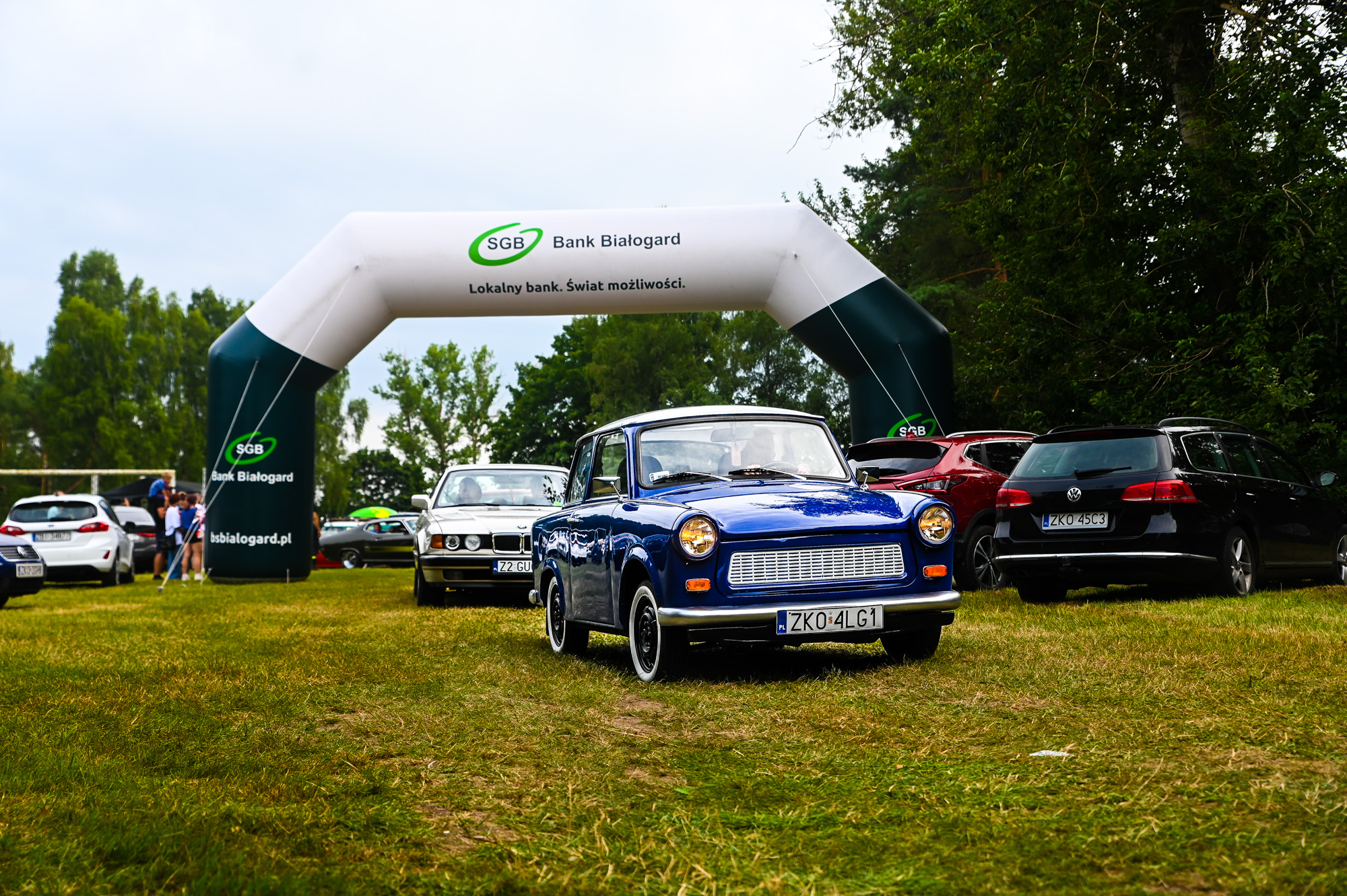 Na zdjęciu widoczny niebieski zabytkowy pojazd marki trabant, w tle r&oacute;żne inne pojazdy zabytkowe oraz balon sponsora Bank Sp&oacute;łdzielczy Białogard