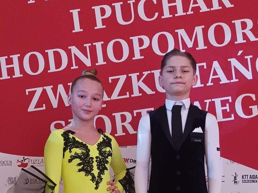  Na zdjęciu Aleksander i Kaja. Kaja ubrana w długą neonową sukienkę w odcieniu żółci z czarnymi dodatkami. Aleksander ubrany w białą koszulę z czarnym krawatem, w czarne spodnie i welurową czarną kamizelkę. Dumnie się prezentują. Na tle widać napis Mistrzostwa i Puchar Zachodniopomorskiego Związku Tańca Sportowego. 