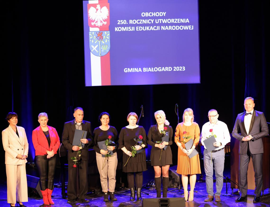 12. Na scenie od lewej stoi Przwodnicząca Rady Gminy, Dyrektor SP w Rogowie, 6 nauczycieli, Wójt Gminy Białogard. Nauczyciele trzymają teczki oraz róże. W tle napis Obchody 250. rocznicy utworzenia Komisji Edukacji Narodowej Gmina Białogard 2023.