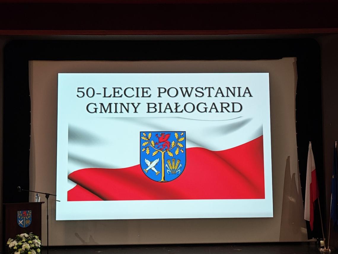 duży, kinowy ekran. Na białym tle widoczny napis 50-lecie powstania Gminy Białogard. Pod napisem biało-czerwona flaga a na jej tle herb Gminy Białogard. Kolory herbu to niebieski, pomarańczowy, biały i czerwony.